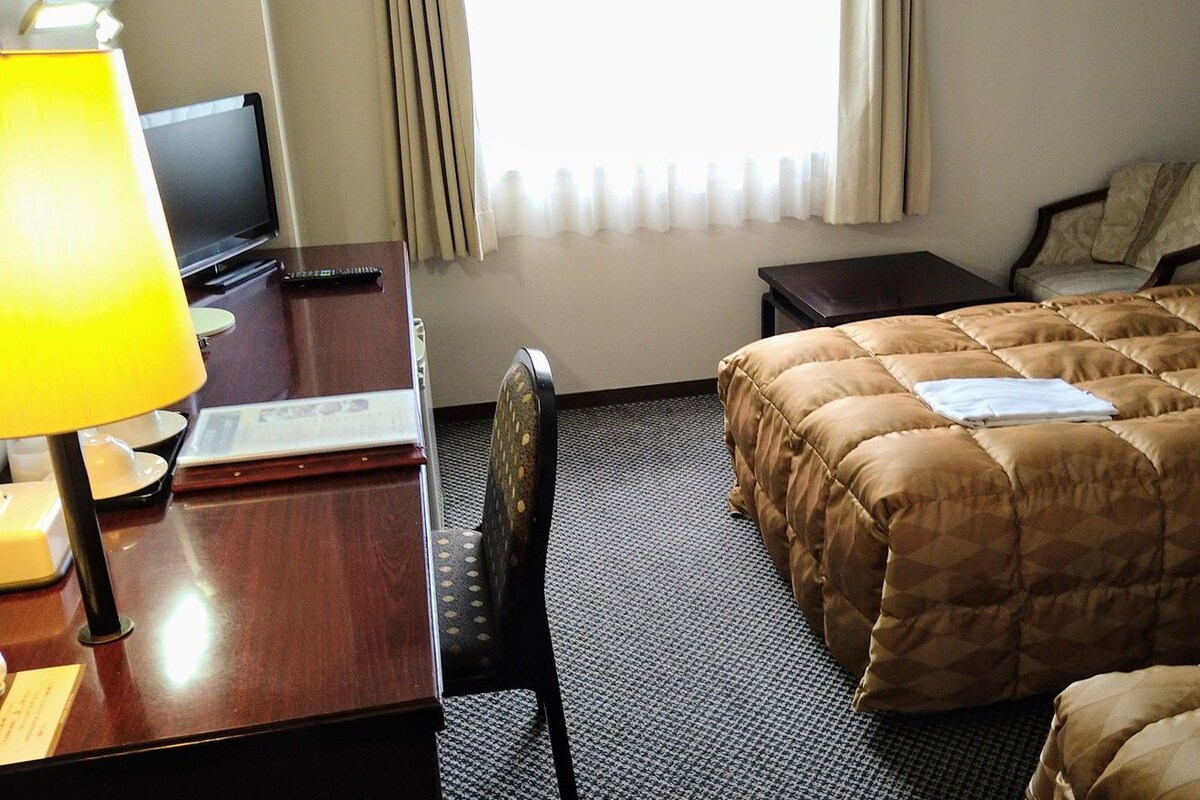 Quarto de hotel com cama, escrivaninha com luminária, cadeira, televisão e janela com cortinas.