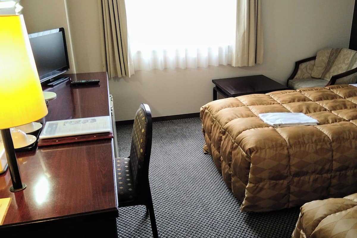 Ein Hotelzimmer mit einem Bett, einem Schreibtisch mit Lampe, einem Sessel, einem Fernseher und einem Fenster mit Vorhängen.