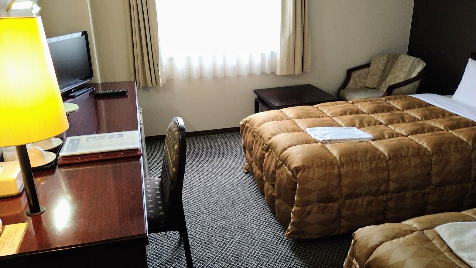 A hotel room with a bed, a desk with a lamp, a chair, a television, and a window with curtains.