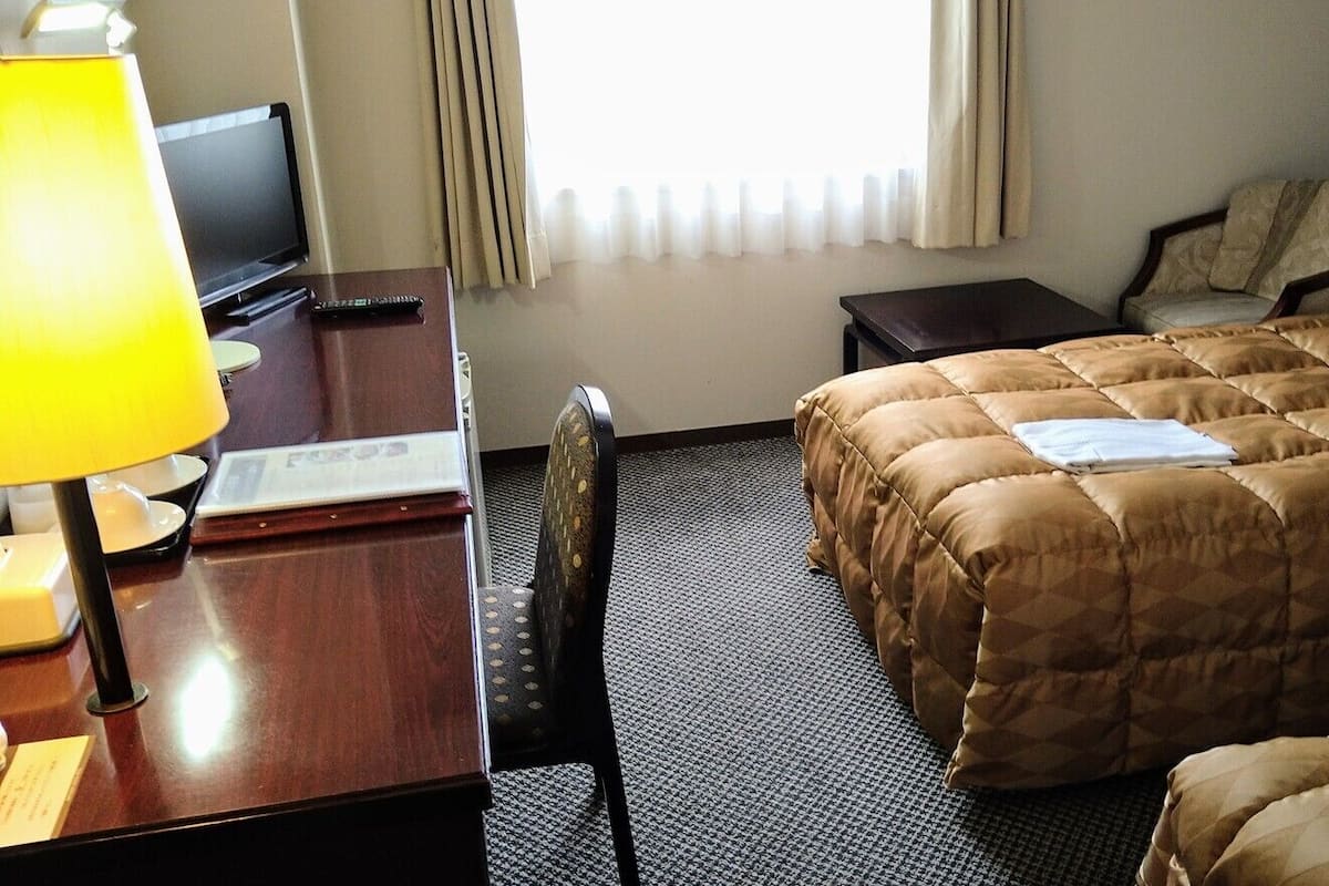 Ein Hotelzimmer mit Bett, Schreibtisch mit Lampe, Sessel, Fernseher und einem Fenster mit Vorhängen.