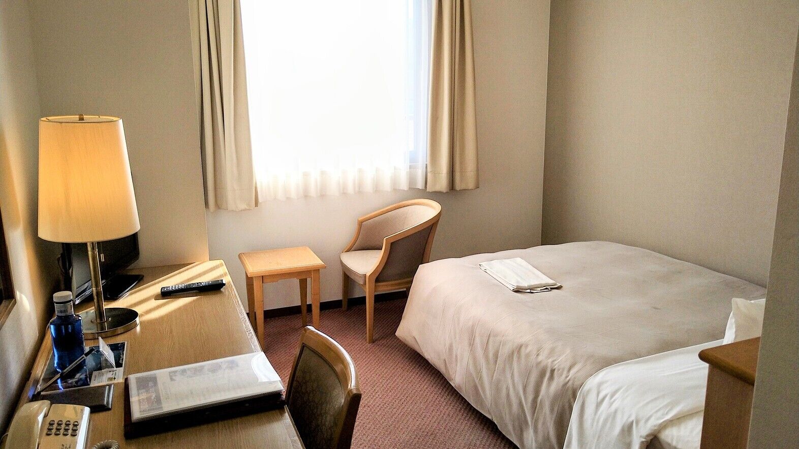 A hotel room with a bed, a desk with a lamp, a chair, a small table, and a window with curtains.