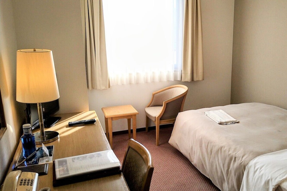 Ein Hotelzimmer mit einem Bett, einem Schreibtisch mit Lampe, einem Sessel, einem kleinen Tisch und einem Fenster mit Vorhängen.