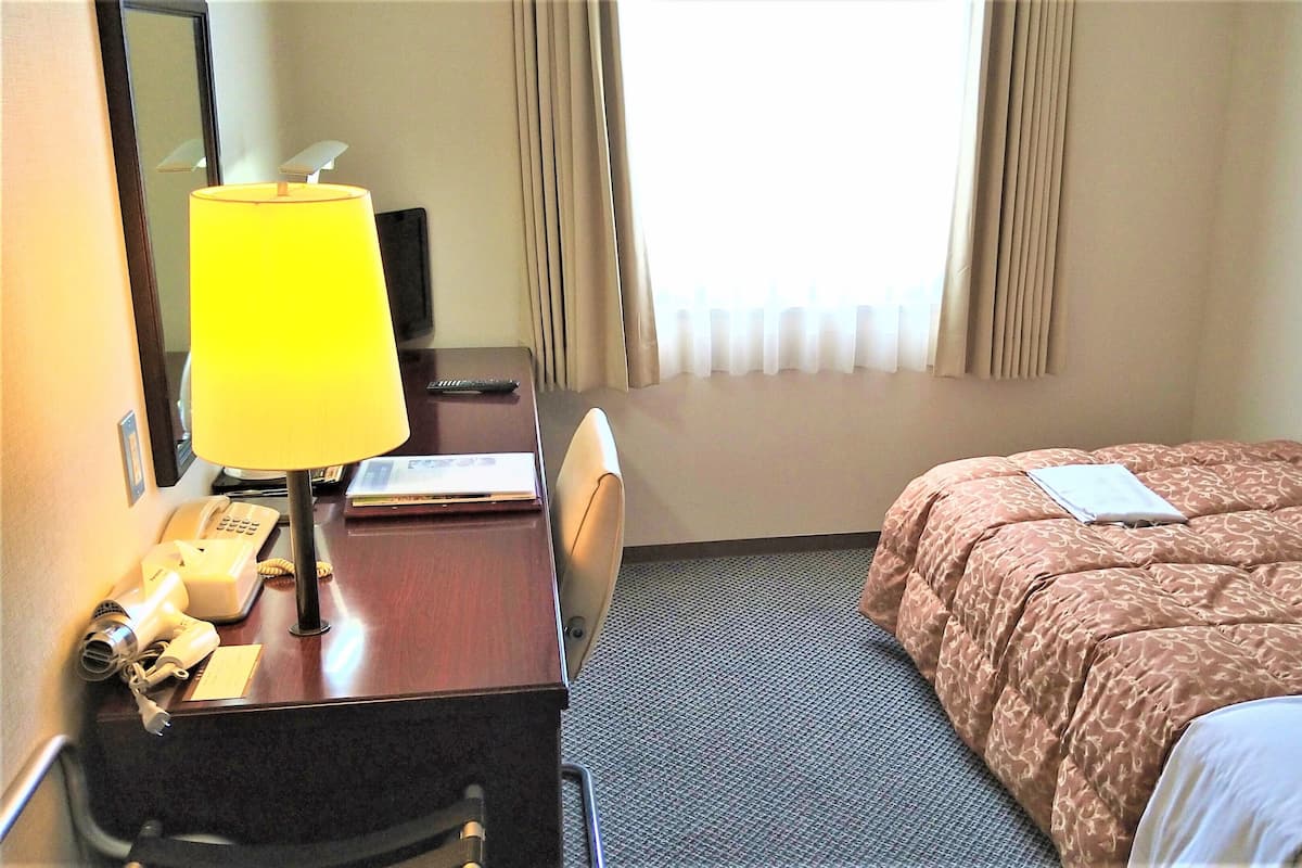 Ein Hotelzimmer mit Bett, Schreibtisch mit Lampe, Telefon und einem Fenster mit Vorhängen.