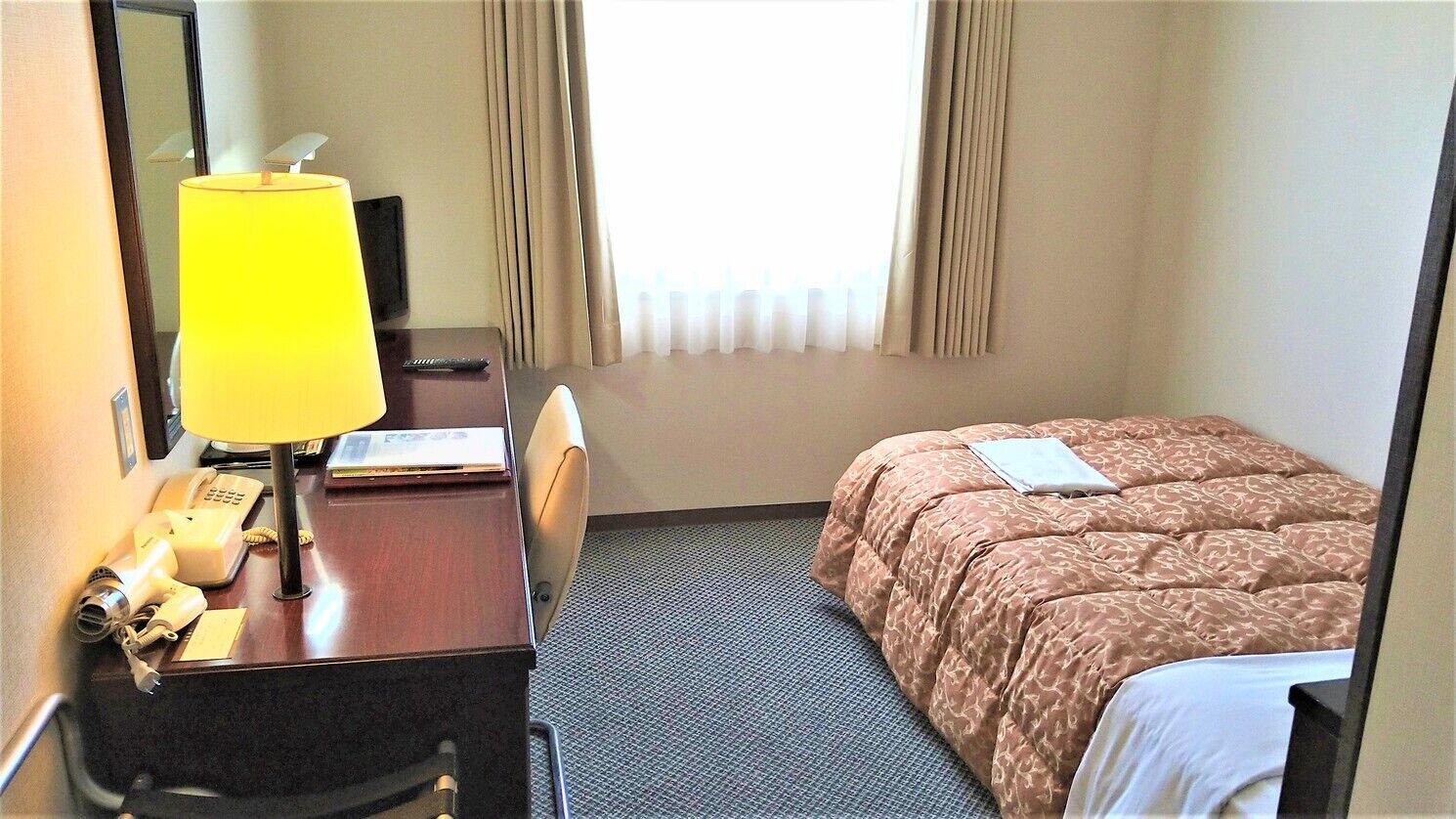 A hotel room with a bed, a desk with a lamp, a telephone, and a window with curtains.