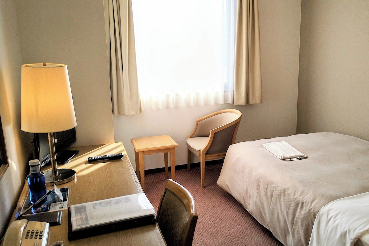 Ein Hotelzimmer mit einem Bett, einem Schreibtisch mit Lampe, einem Sessel, einem Telefon und einem Fenster mit Vorhängen.