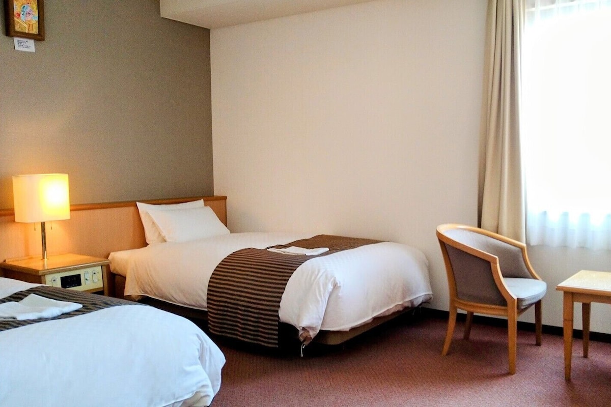 Ein Hotelzimmer mit zwei Betten, einem Schreibtisch, einem Stuhl, einer Lampe und einem Vorhangfenster.