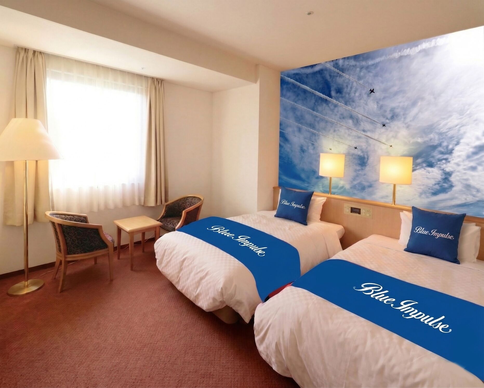 A hotel room with two beds, a desk, a chair, a window with curtains, and a wall mural of a sky with clouds and airplanes.