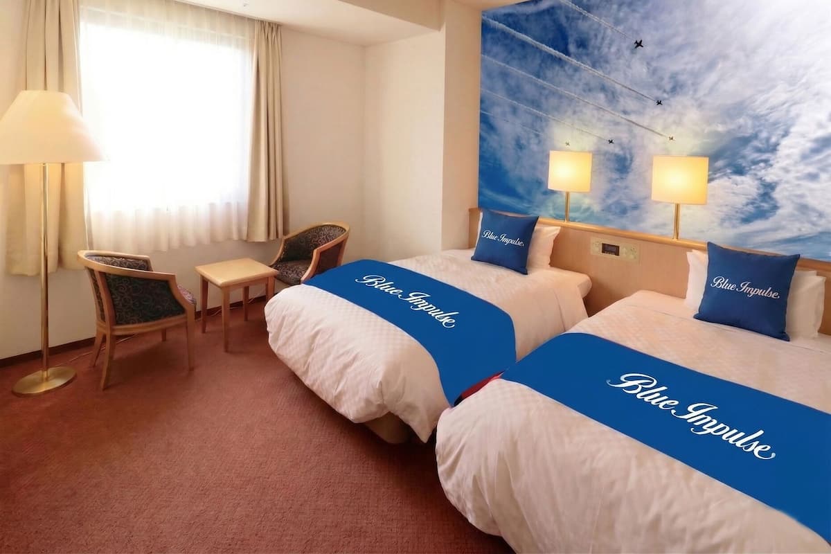 Ein Hotelzimmer mit zwei Betten, einem Schreibtisch, einem Stuhl, einem Fenster mit Vorhängen und einem Wandbild mit Himmel, Wolken und Flugzeugen.