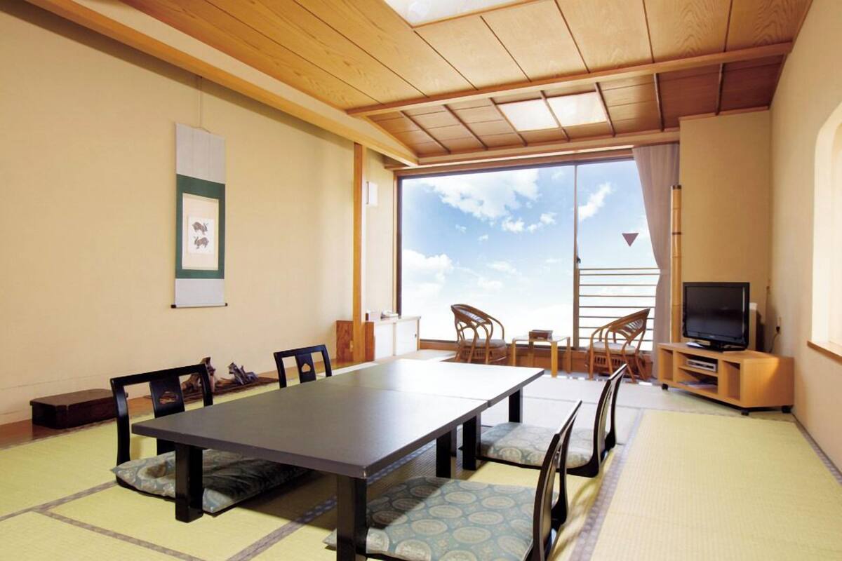 Ein Zimmer mit Tatami-Boden, einem langen Tisch, Stühlen, einem Fernseher und einem Balkon mit Blick.