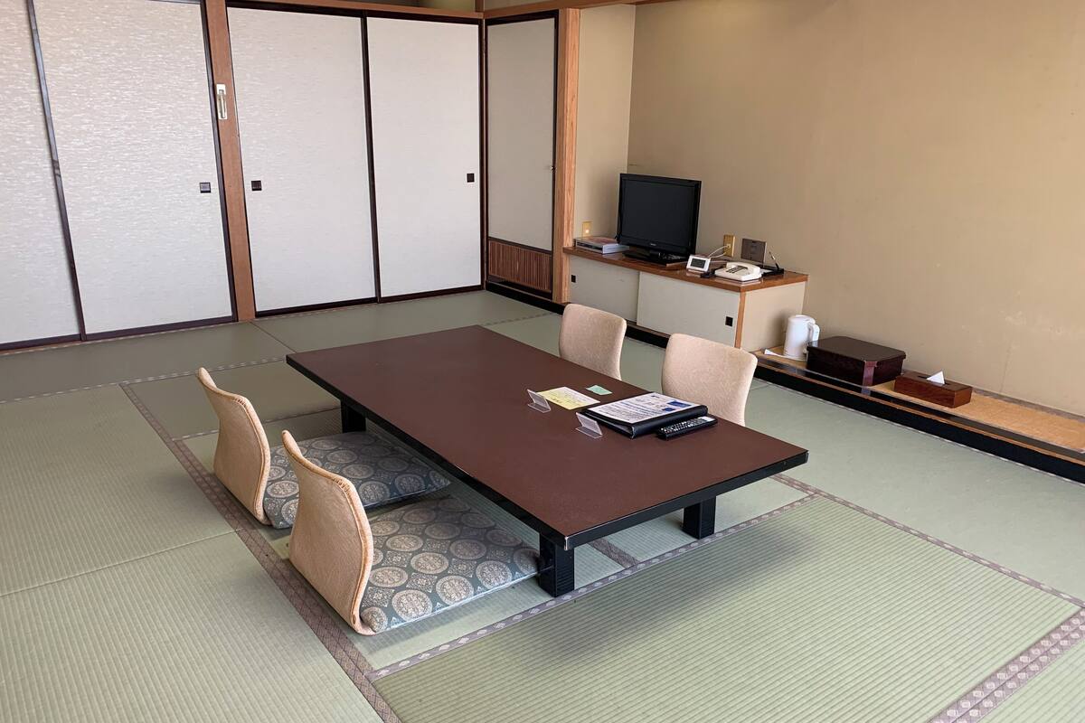 Ein traditioneller japanischer Raum mit Tatami-Boden, einem niedrigen Tisch mit Kissen, einem Fernseher und einem kleinen Schrank mit Telefon und weiteren Gegenständen.
