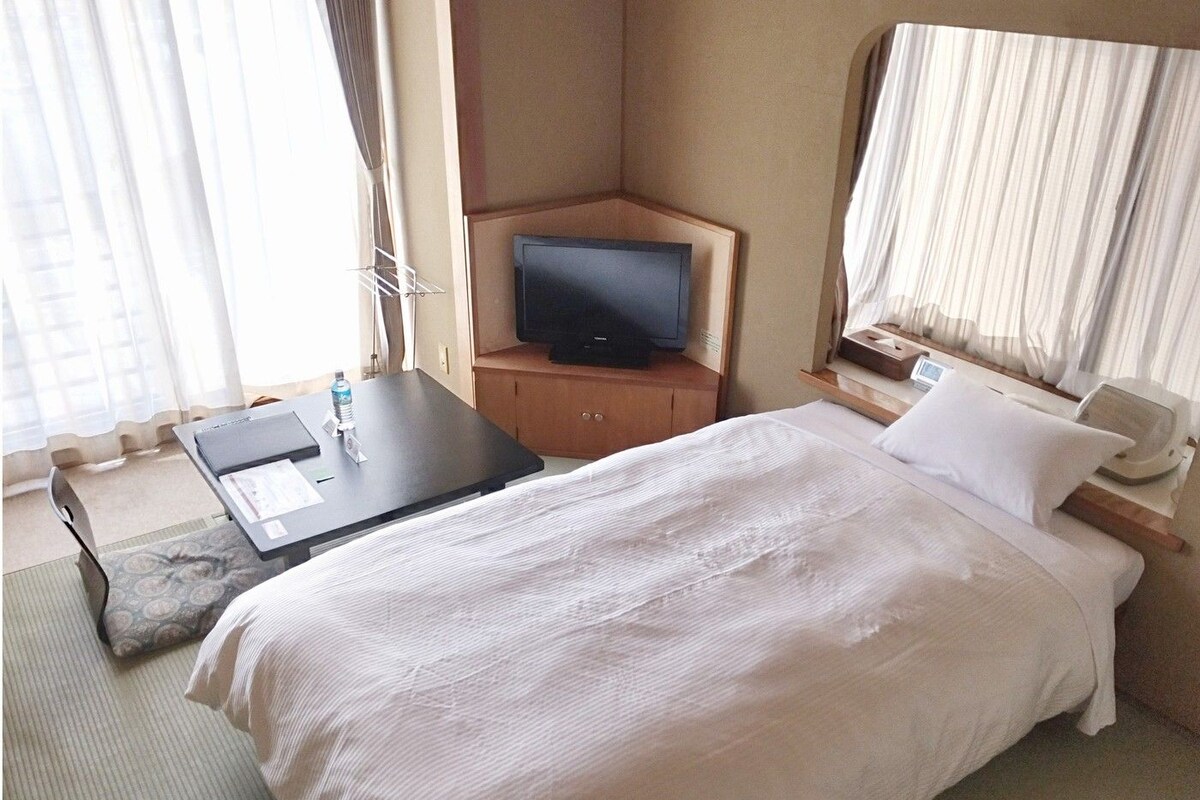 A hotel room with a bed, a desk with a television, a bedside table with a lamp, and a window with curtains.