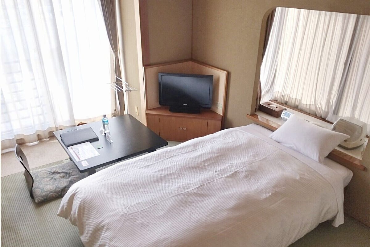 Ein Hotelzimmer mit Bett, Schreibtisch mit Fernseher, Nachttisch mit Lampe und Fenster mit Vorhängen.