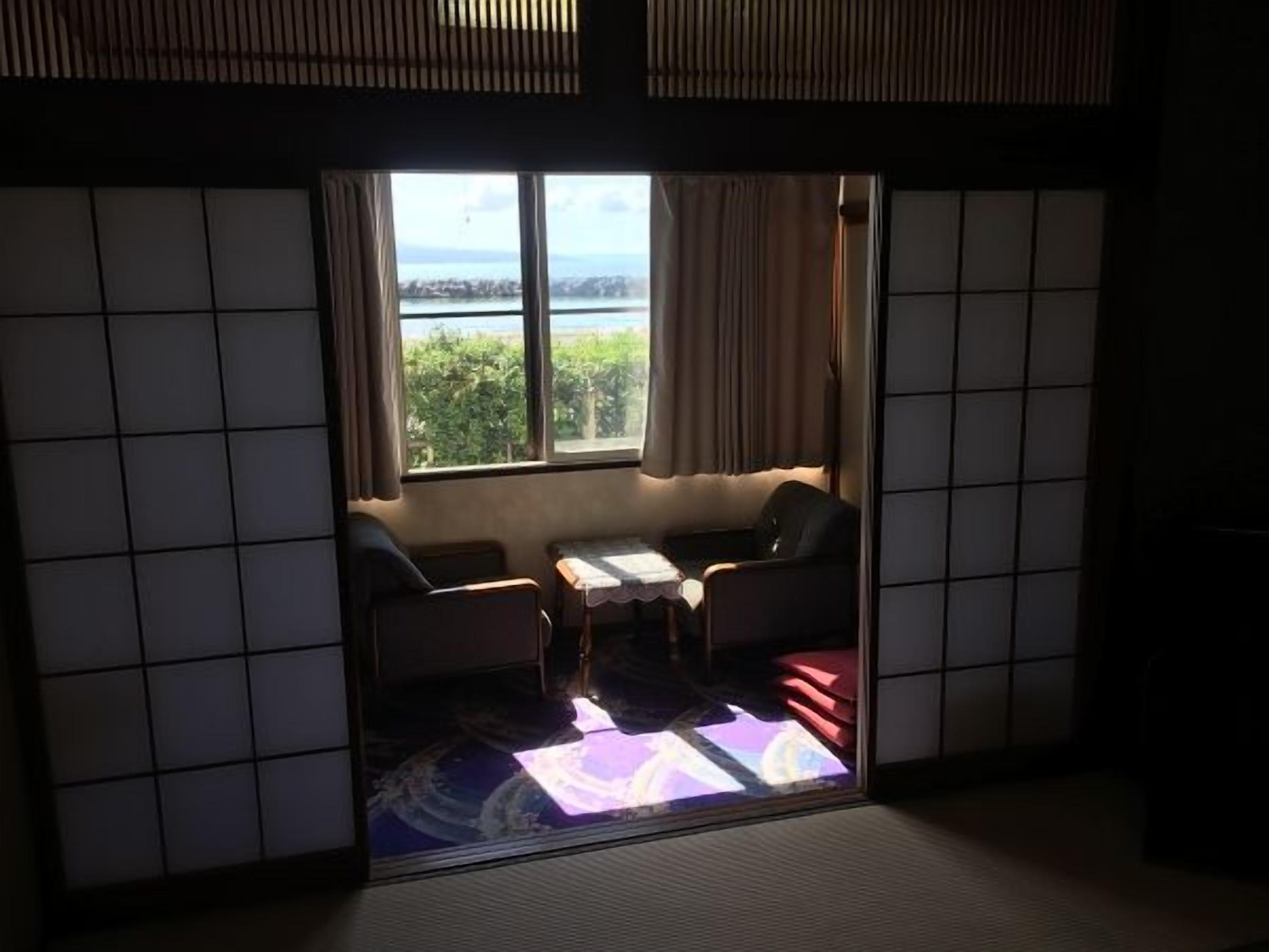 A room with tatami flooring, a low table, a sofa, and a view of a body of water through a window.