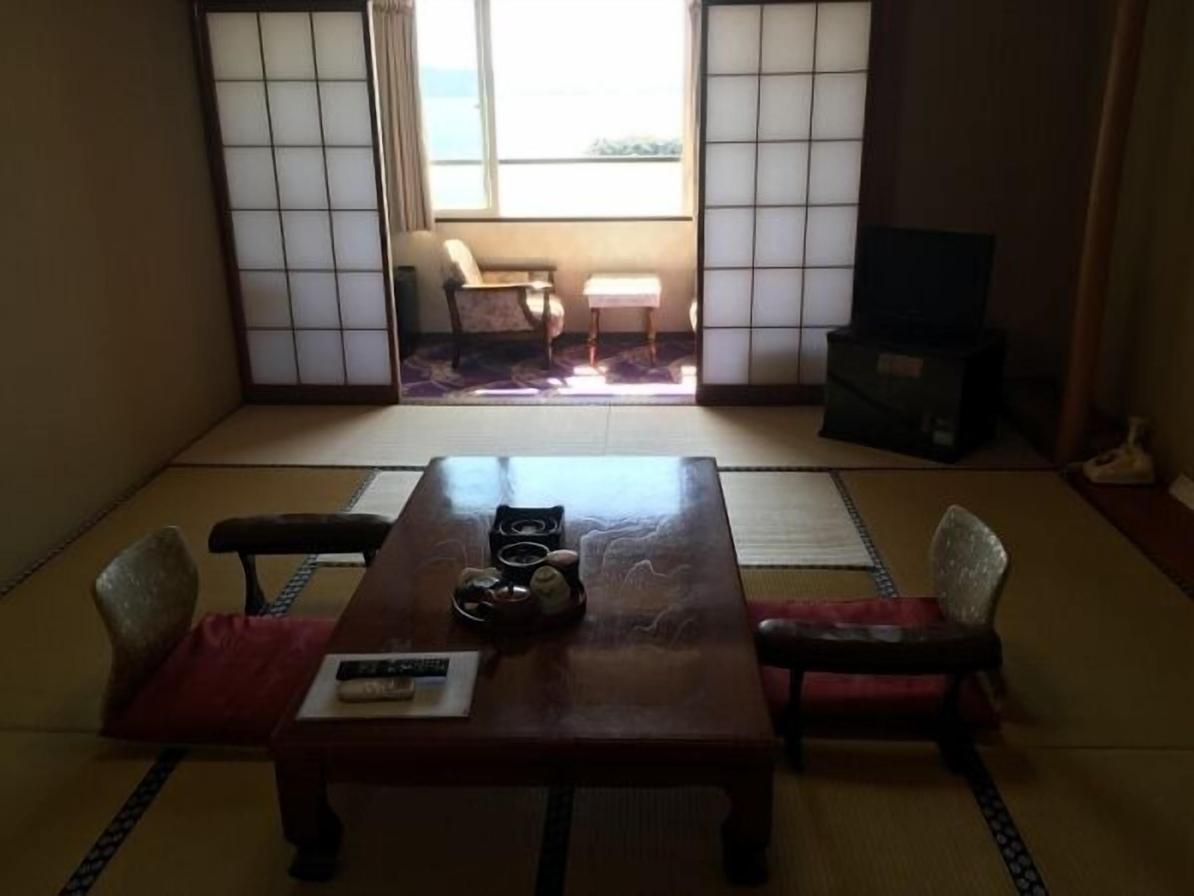 Ein traditioneller japanischer Raum mit Tatami-Boden, einem niedrigen Tisch mit Kissen, einem Fernseher und Schiebetüren.