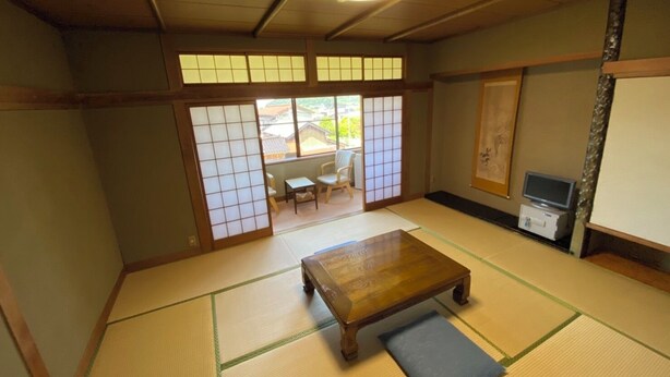 Ein traditioneller japanischer Raum mit Tatami-Böden, einem Holztisch, einer Futonmatte auf dem Boden, einer Schiebetür und einem Fernseher auf einem Standfuß.