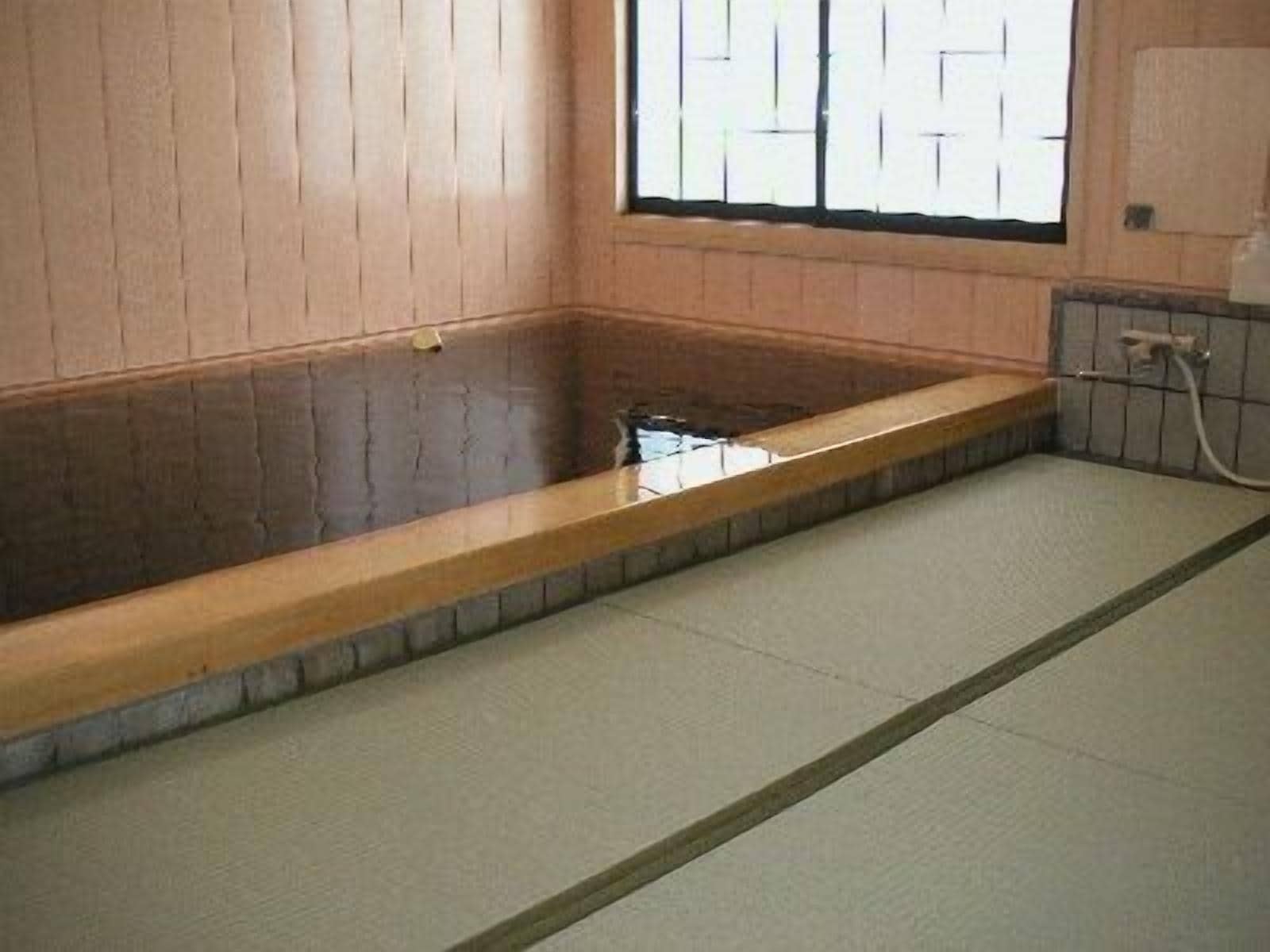 A traditional Japanese onsen with a wooden bath, tatami flooring, and a view through a shoji screen.