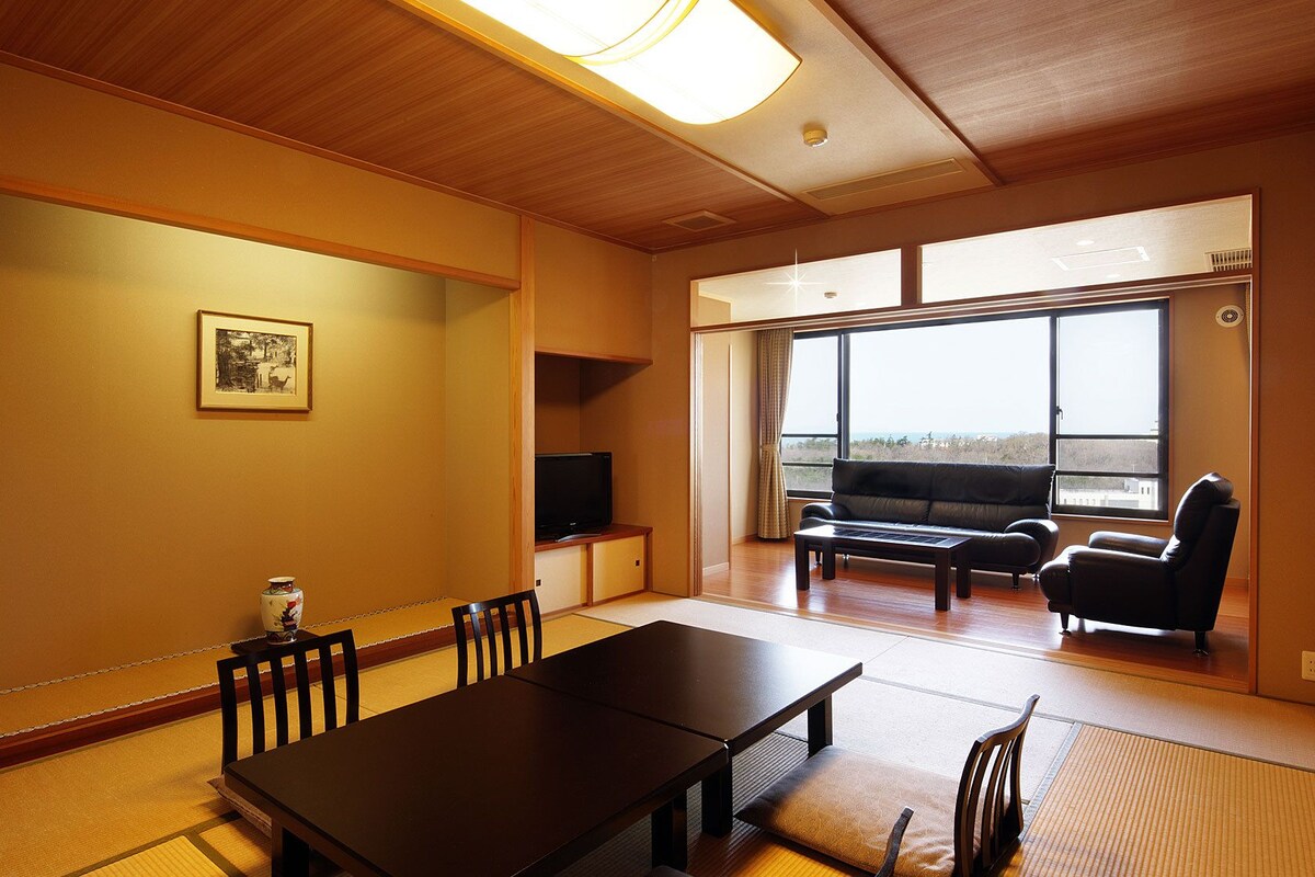 A room with tatami flooring, a dining table, chairs, a sofa, a TV, and large windows with a view.