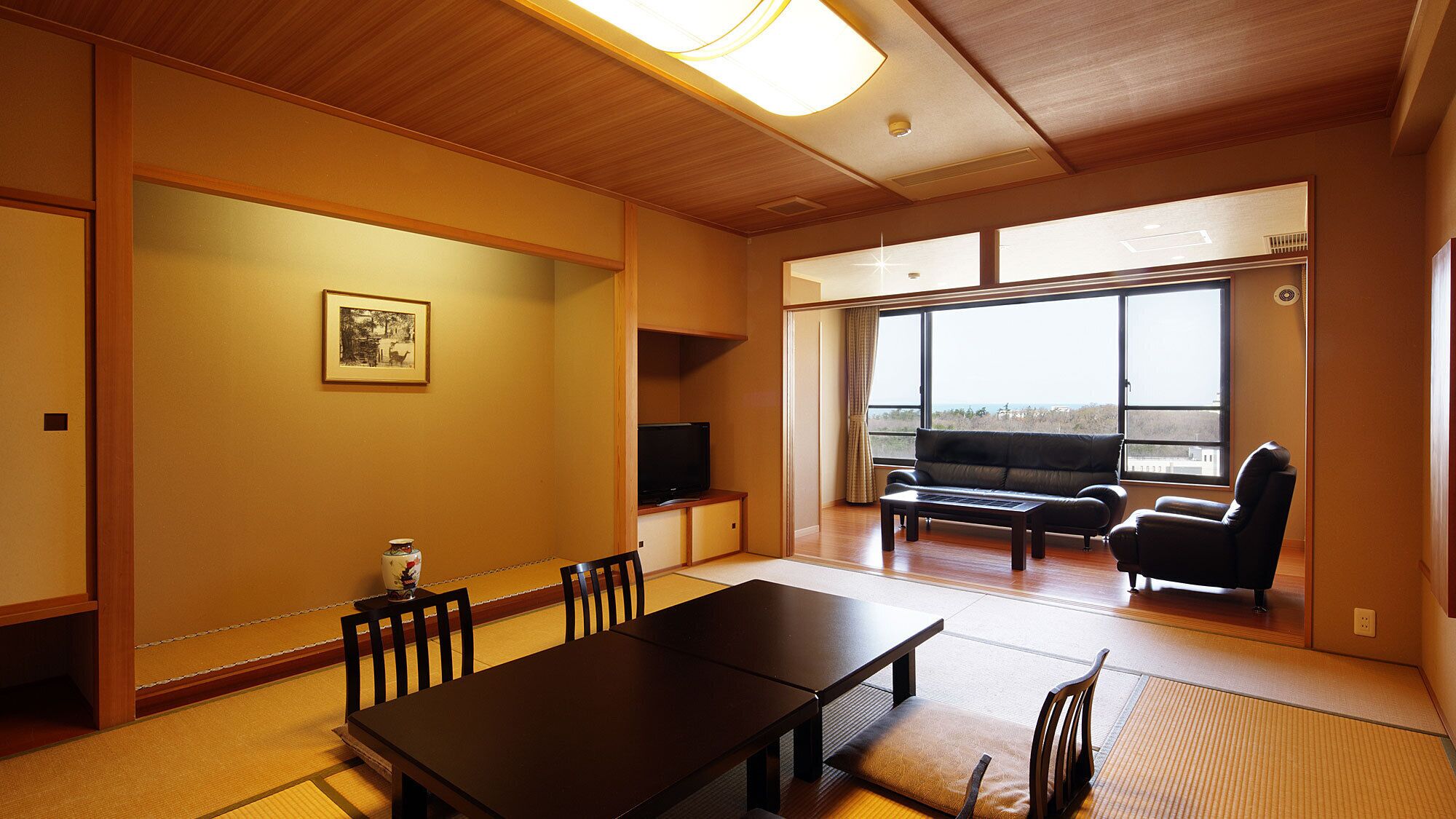 A room with tatami flooring, a dining table, chairs, a sofa, a TV, and large windows with a view.