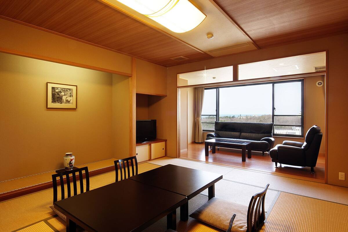 A room with tatami flooring, a dining table, chairs, a sofa, a TV, and large windows with a view.