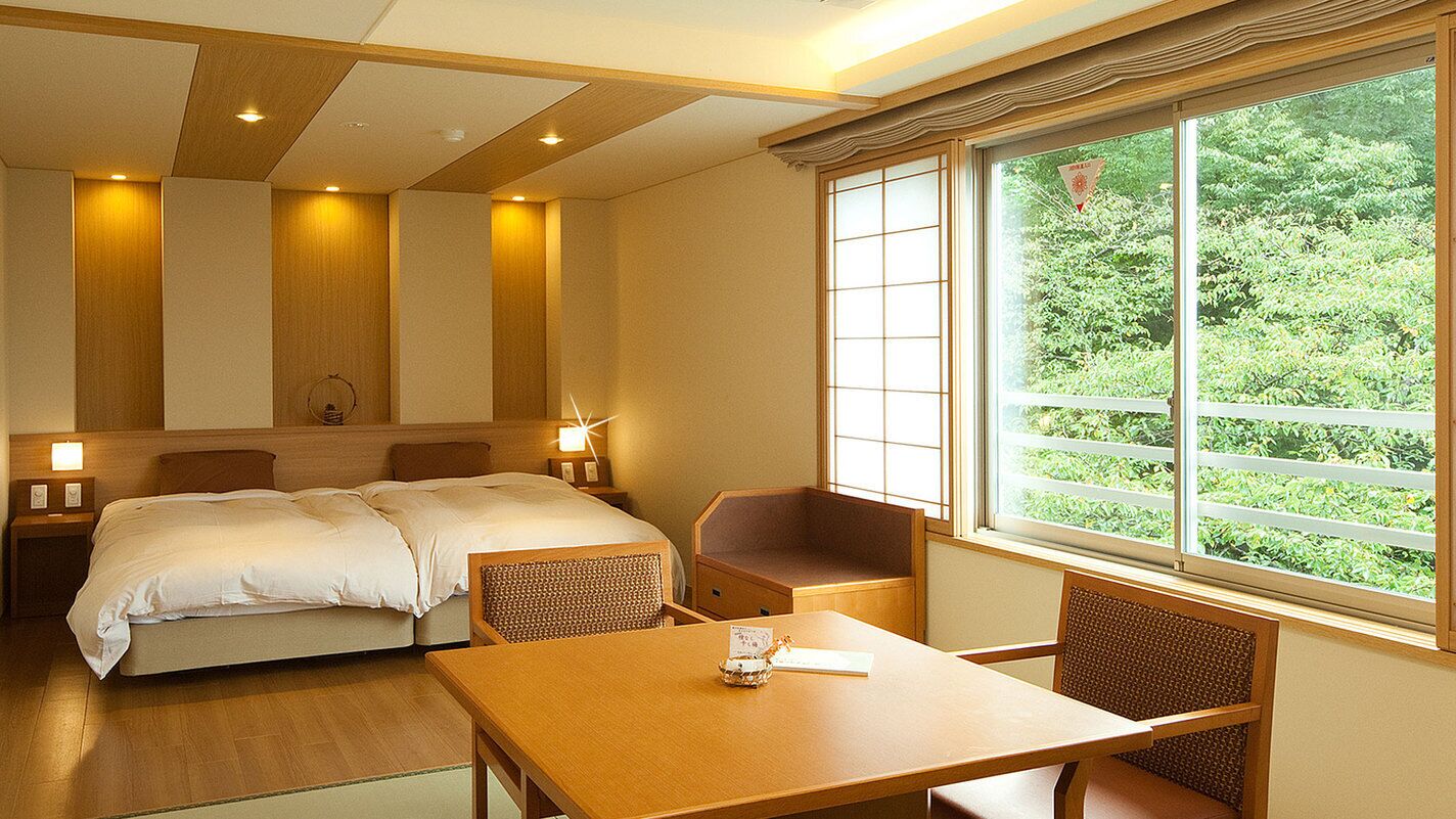 A hotel room with a bed, a table, chairs, a window with a view of greenery, and a wooden ceiling.