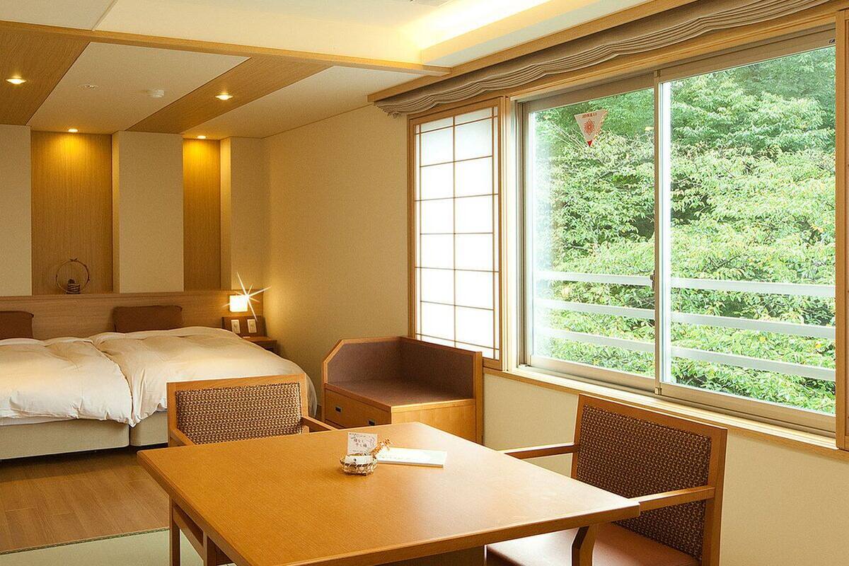 A hotel room with a bed, a table, chairs, a window with a view of greenery, and a wooden ceiling.