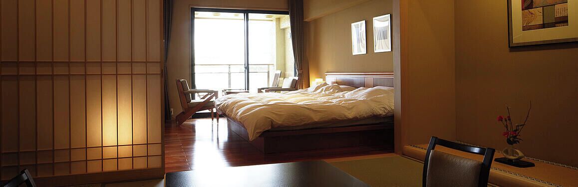 A hotel room with a bed, a chair, a table, and a sliding door leading to a balcony.