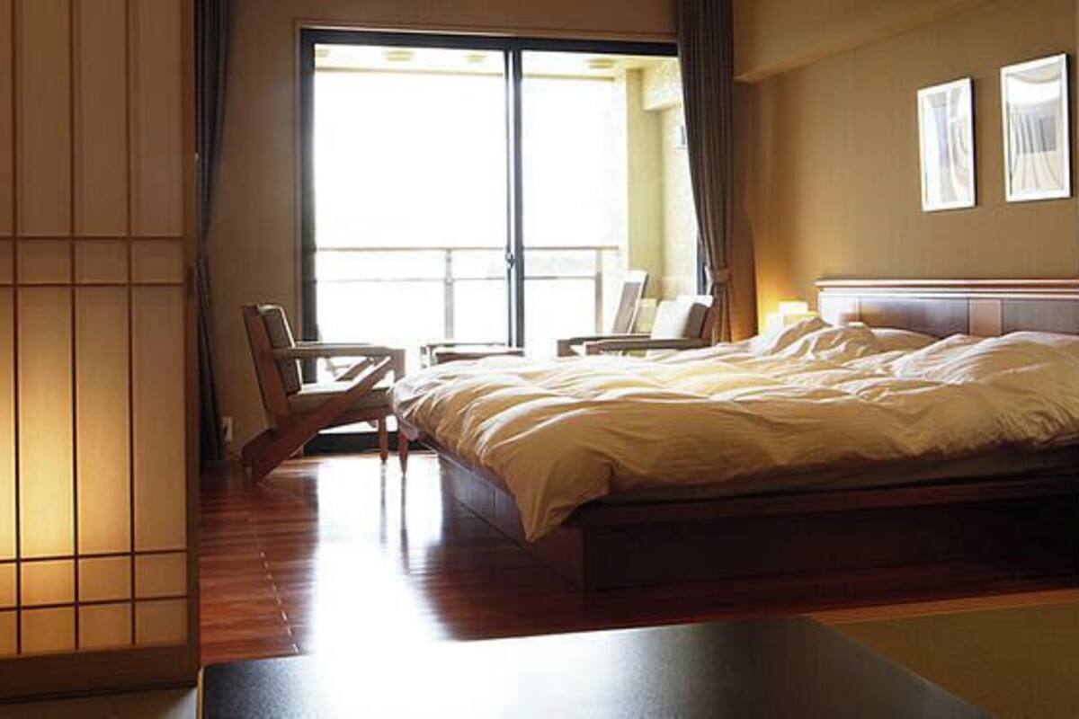 A hotel room with a bed, a chair, a table, and a sliding door leading to a balcony.