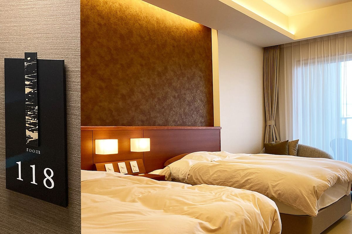 A hotel room with two beds, a large window with curtains, and a wall-mounted sign indicating room number 118.