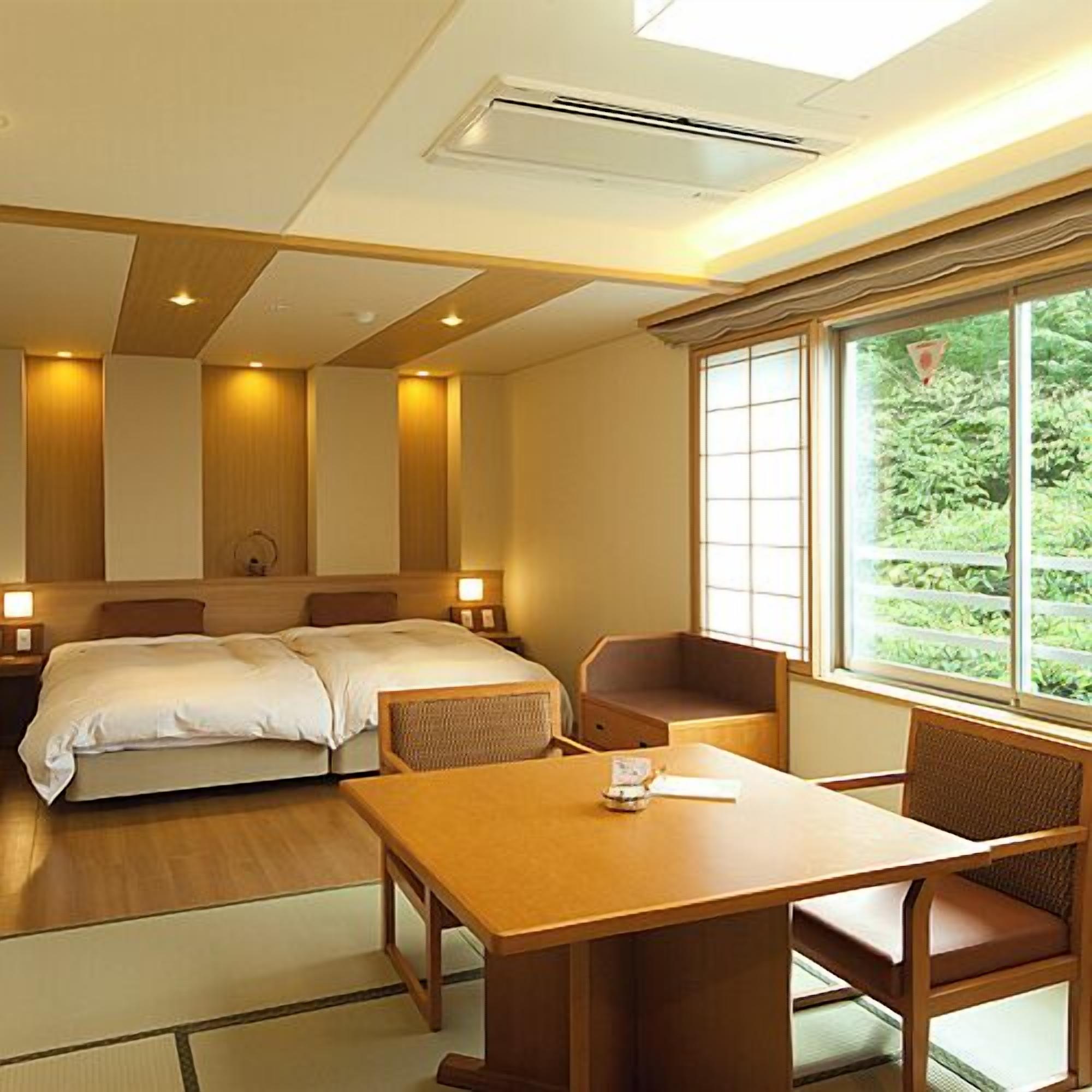 A hotel room with two beds, a dining table, and a window with a view of greenery.
