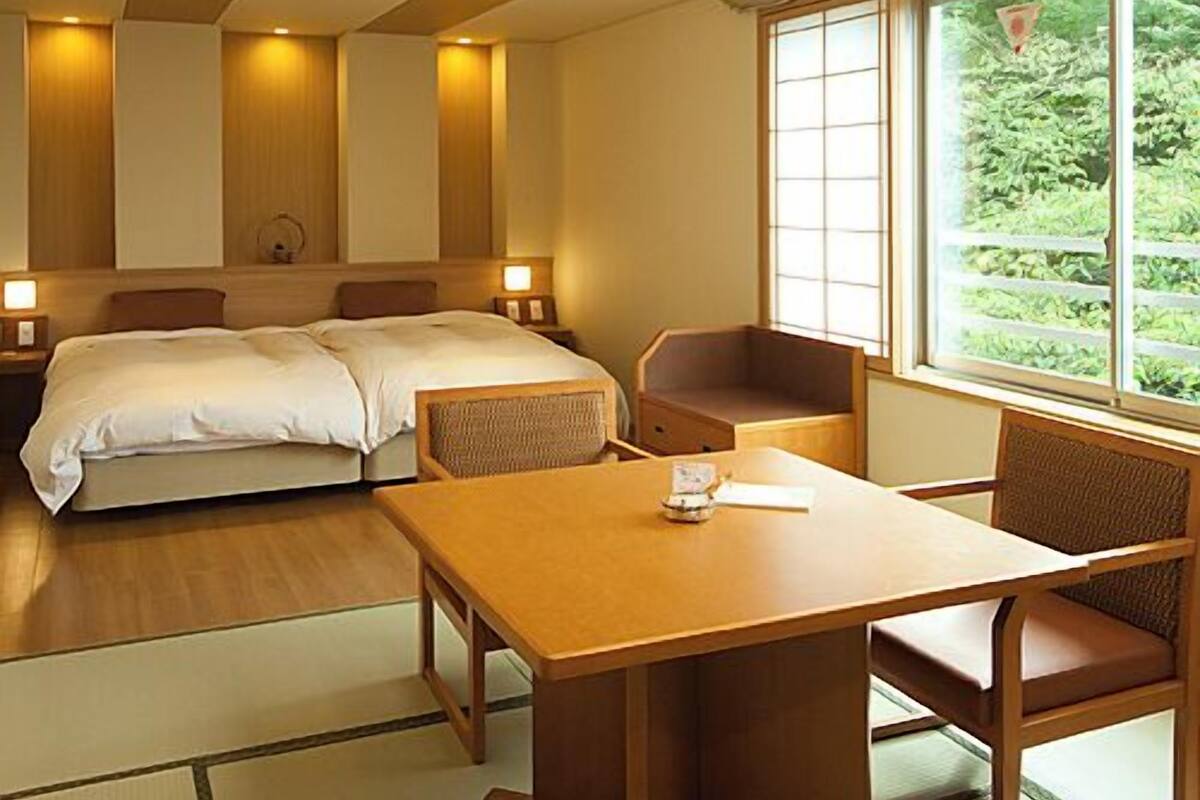 A hotel room with two beds, a dining table, and a window with a view of greenery.