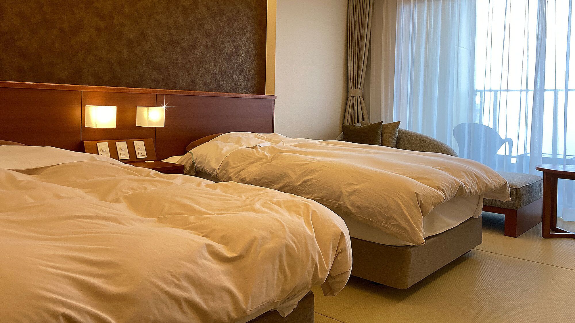 A hotel room with two beds, a nightstand with lamps, a chair, and a view of the outdoors.