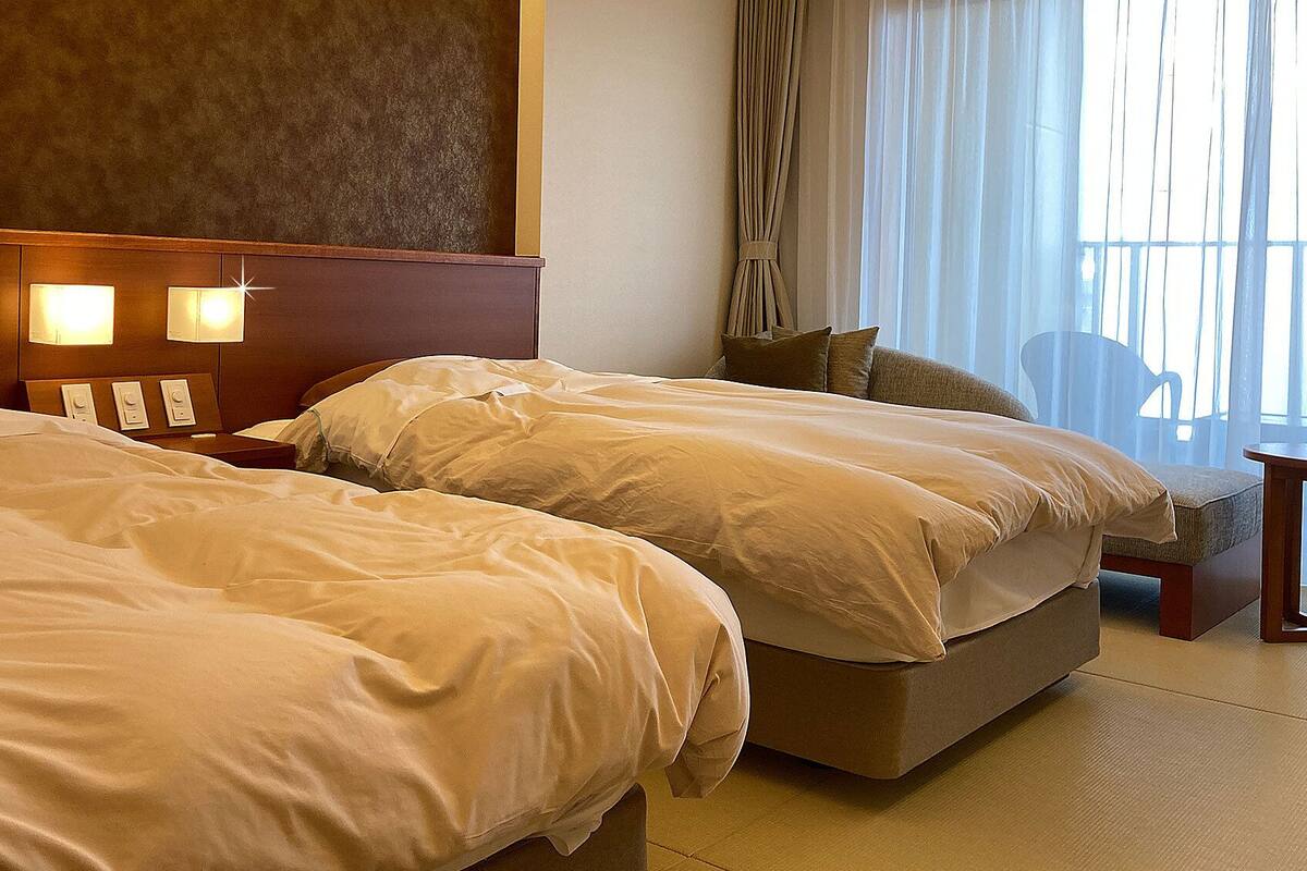 A hotel room with two beds, a nightstand with lamps, a chair, and a view of the outdoors.