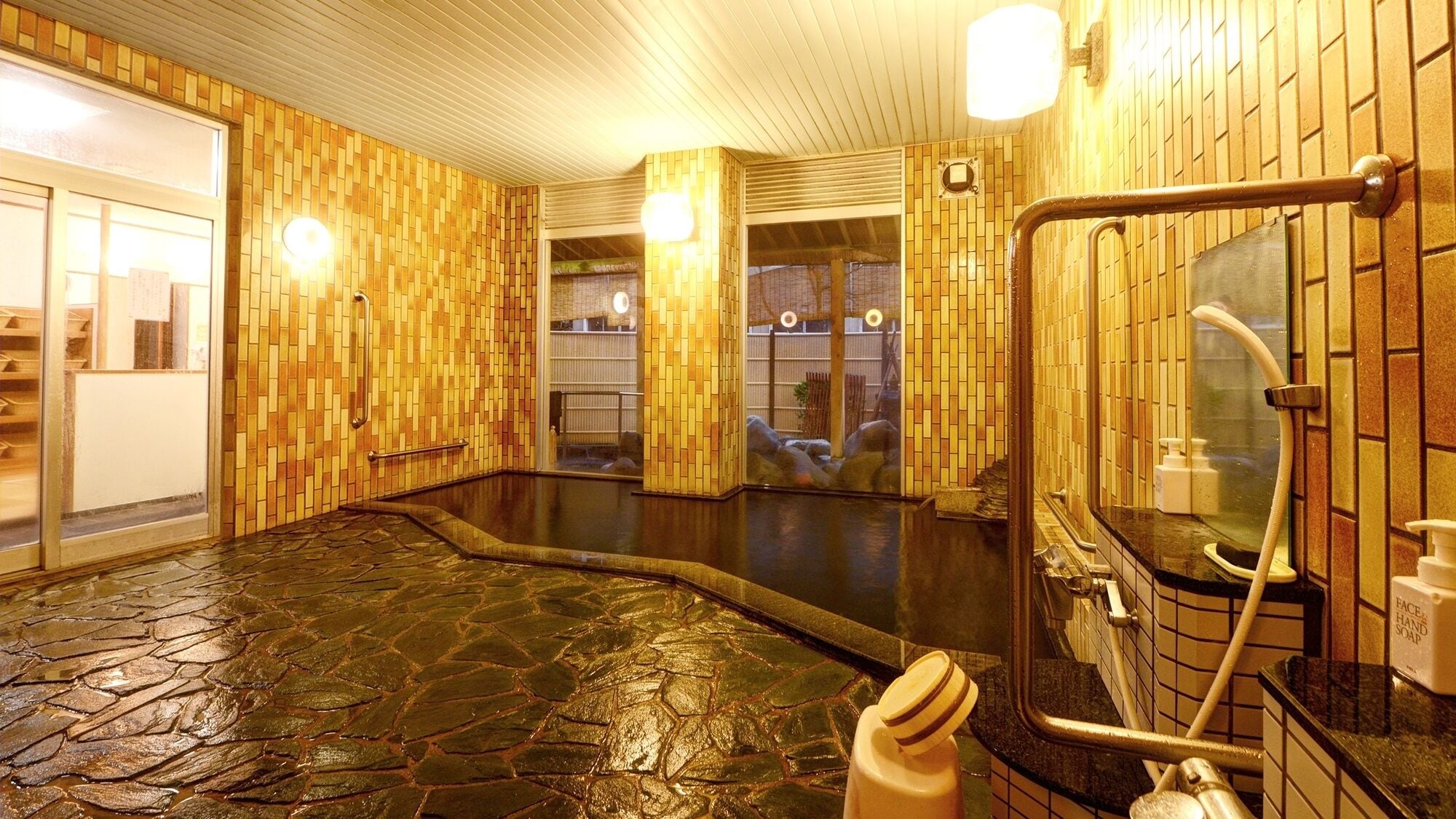 A traditional Japanese onsen with a stone floor, wooden walls, and a glass-enclosed shower area.