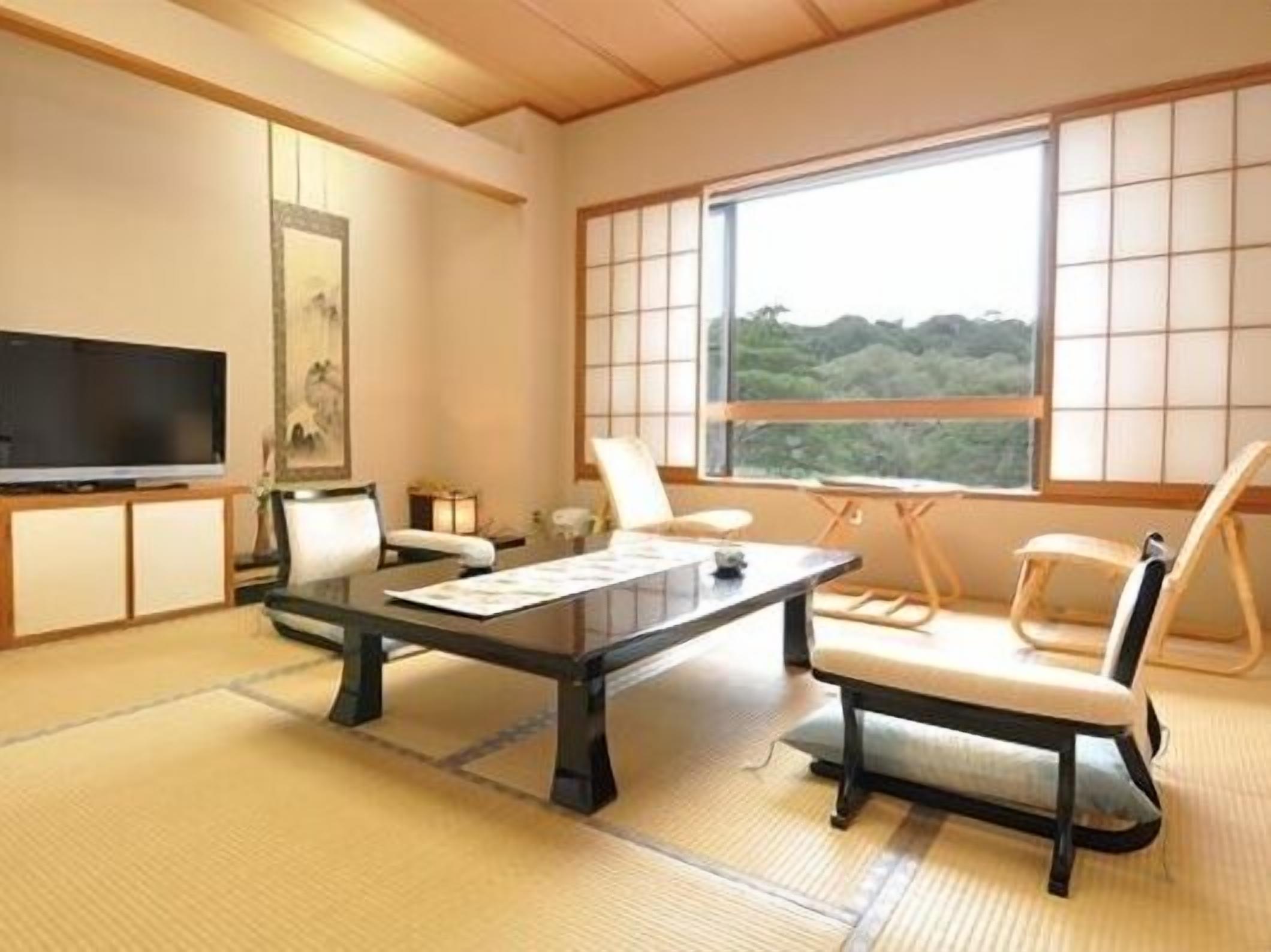 Une pièce de style japonais traditionnel, avec un sol recouvert de tatamis, une télévision, un bureau avec des papiers et une vue sur une forêt, visible à travers de grandes fenêtres.