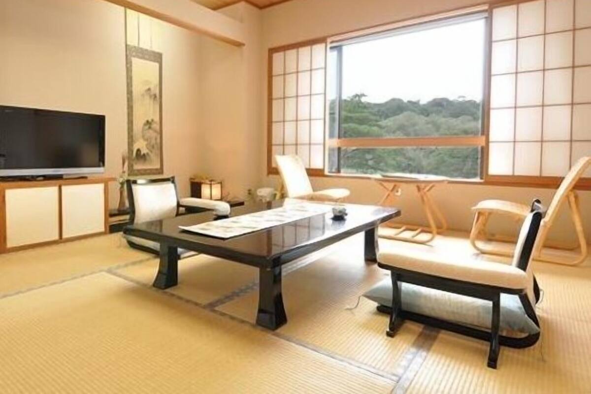 A traditional Japanese-style room with tatami flooring, a television, a desk with papers, and a view of a forest through large windows.