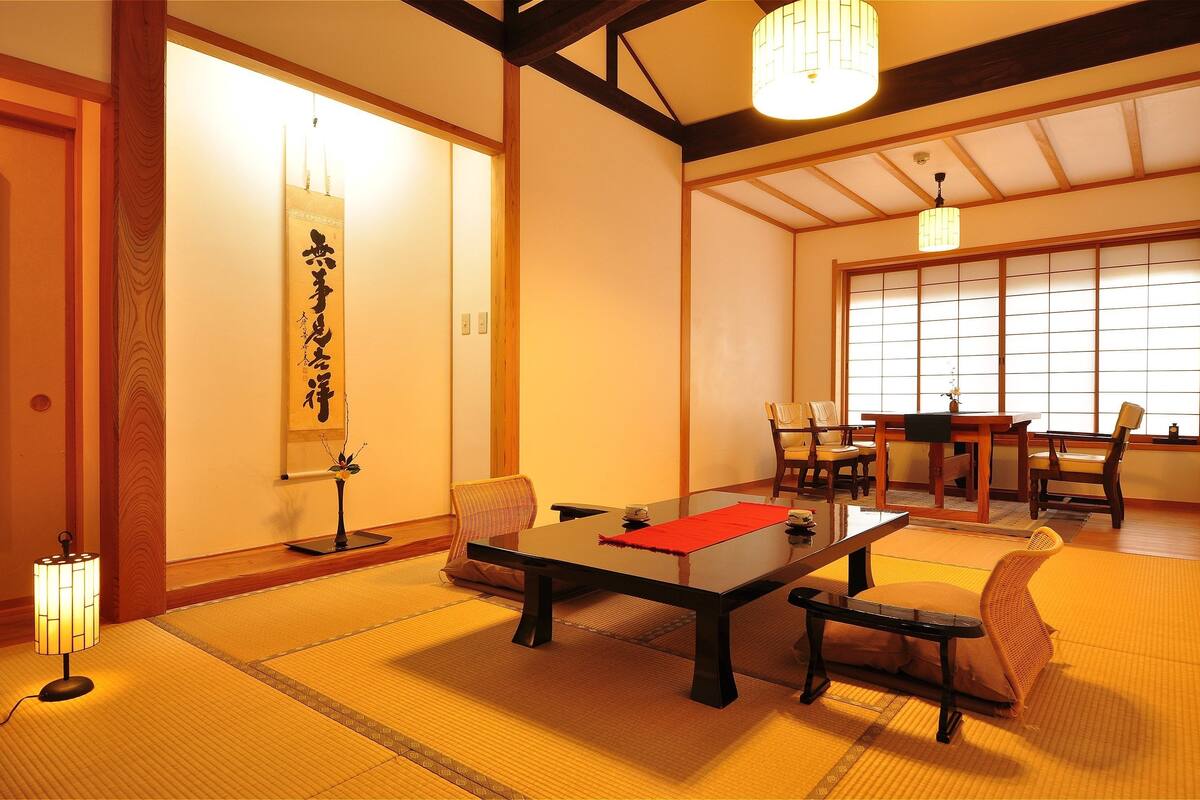 A traditional Japanese room with tatami flooring, a low table, and chairs. There is a hanging scroll on the wall and a lamp on the floor.