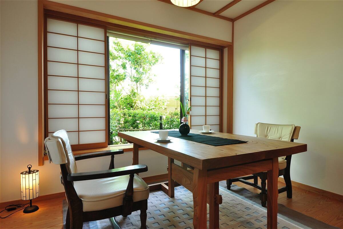 A traditional Japanese room with tatami flooring, a wooden table, two chairs, a sliding shoji screen, and a small table lamp.