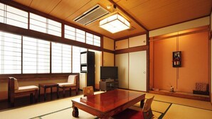 Ein traditioneller japanischer Raum mit Tatami-Boden, einem niedrigen Tisch, Stühlen, einem Fernseher und Schiebetüren.