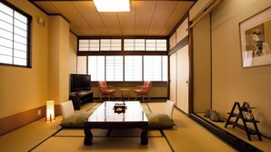Ein traditioneller japanischer Raum mit Tatami-Boden, einem niedrigen Tisch, Stühlen, einem Fernseher und einer Schiebetür.