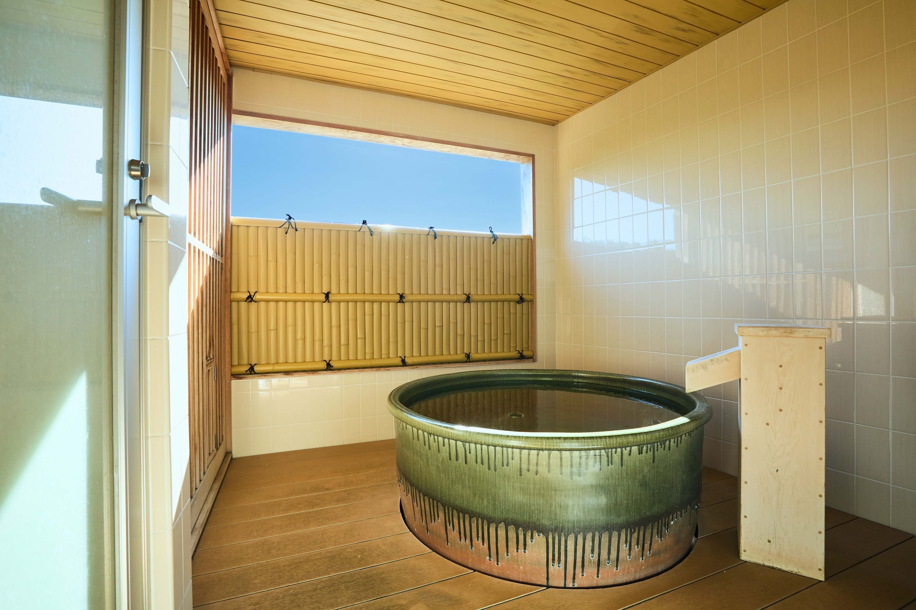 Una habitación con techo de madera, un ventanal con persianas de bambú, una bañera de vidrio y paredes con azulejos.