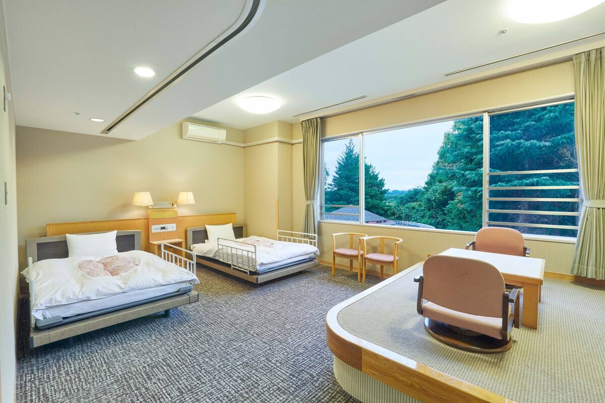 Une chambre d’hôtel équipée d’un lit, d’un bureau, de deux chaises, d’une fenêtre donnant sur la verdure et d’un appareil de climatisation.