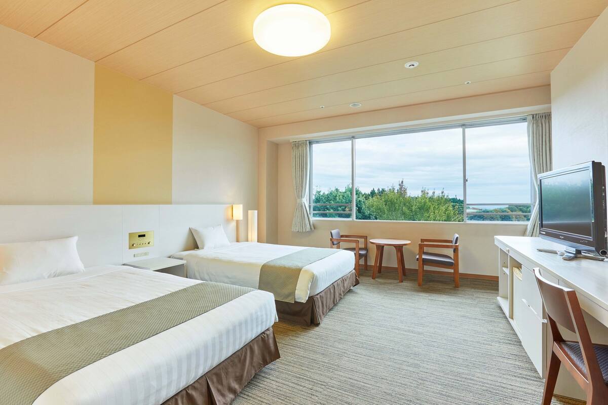 Une chambre d’hôtel avec deux lits, un bureau, une chaise, une télévision et une grande fenêtre donnant sur un paysage verdoyant.