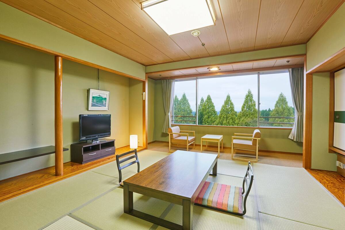 Une pièce de style japonais traditionnel, avec un sol recouvert de tatamis, une petite table en bois, un téléviseur et une vue sur les arbres par la fenêtre.