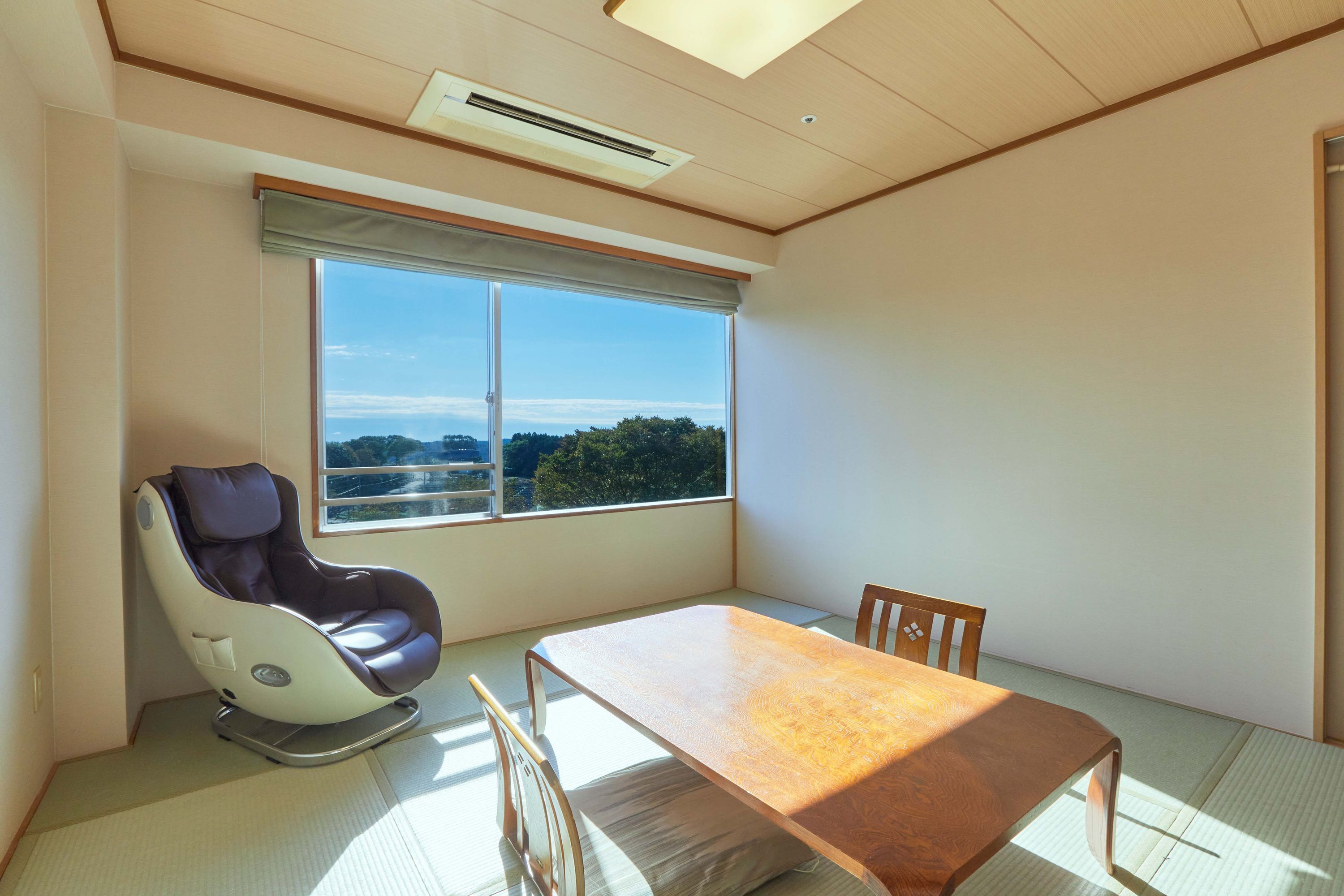 A room with a massage chair, a wooden table, and a large window with a view of trees and hills.