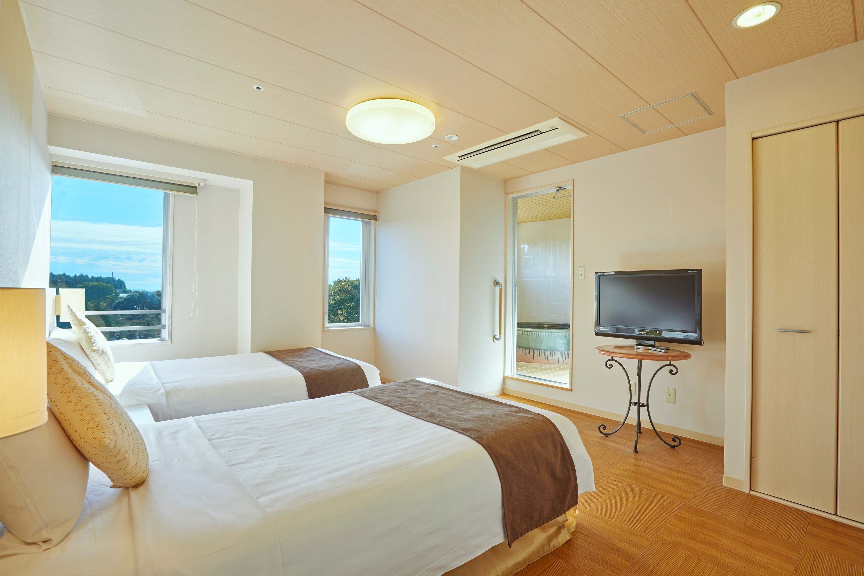 A hotel room with two beds, a TV on a small table, and a large window offering a view of the outdoors.