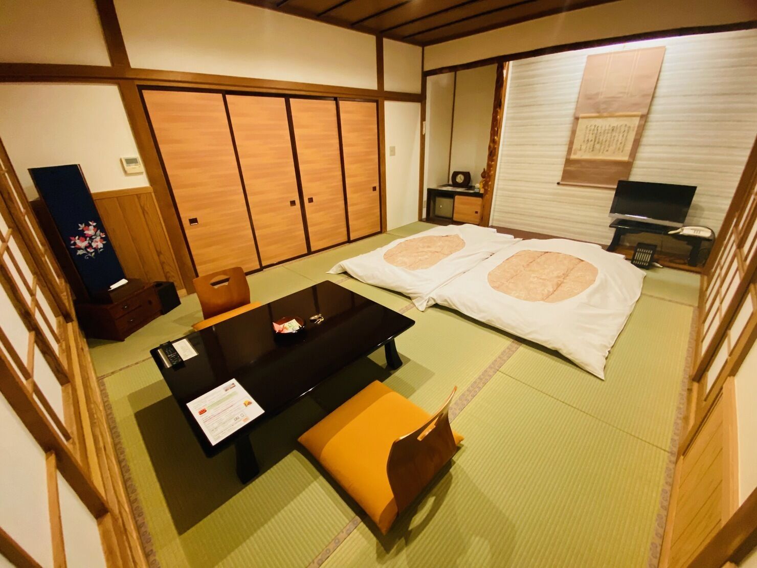 Een traditionele Japanse kamer met tatami vloeren, een futonbed, een lage tafel met een menukaart, een houten kast en een televisie.