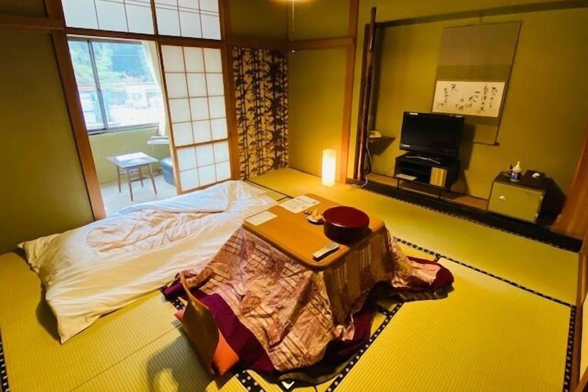A traditional Japanese-style room with tatami flooring, a bed with white bedding, a small table with a red container, a television, and a sliding door leading to a balcony.