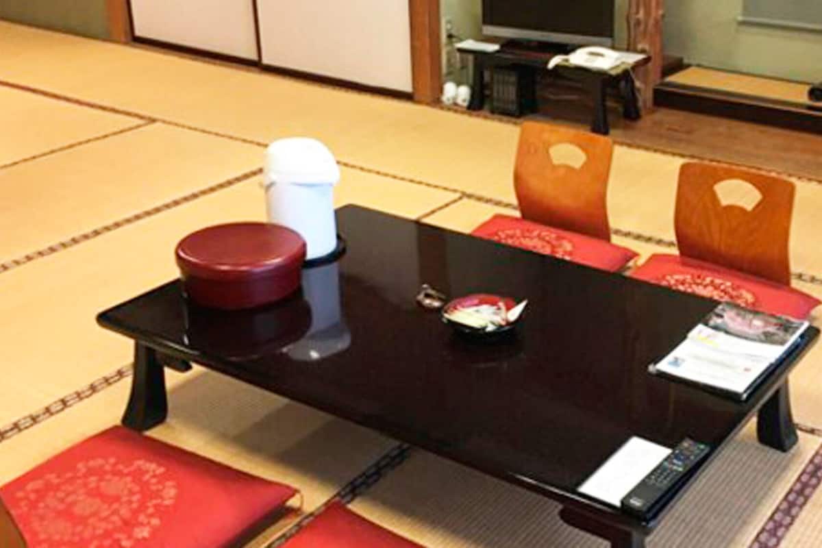 A traditional tatami mat room with a low table, two red cushions, a white container, and a remote control on the table.
