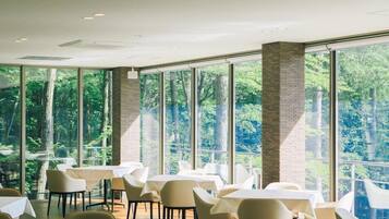 Ein Essbereich mit Tischen und Stühlen, großen Fenstern und Blick auf Bäume.