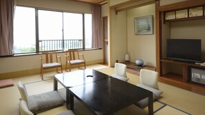 Una habitación japonesa tradicional con suelo de tatami, una mesa baja, sillas, un televisor y un ventanal con cortinas.